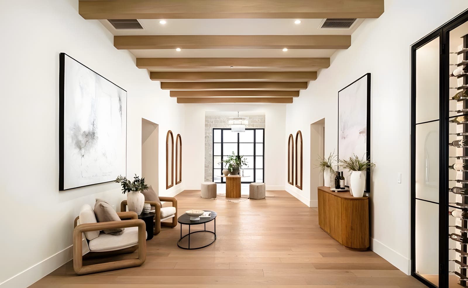 Entry gallery with exposed wood ceiling beams, arched wall niches, white oak hardwood floors, iron-framed glass entry door, and a wine storage wall at the far end